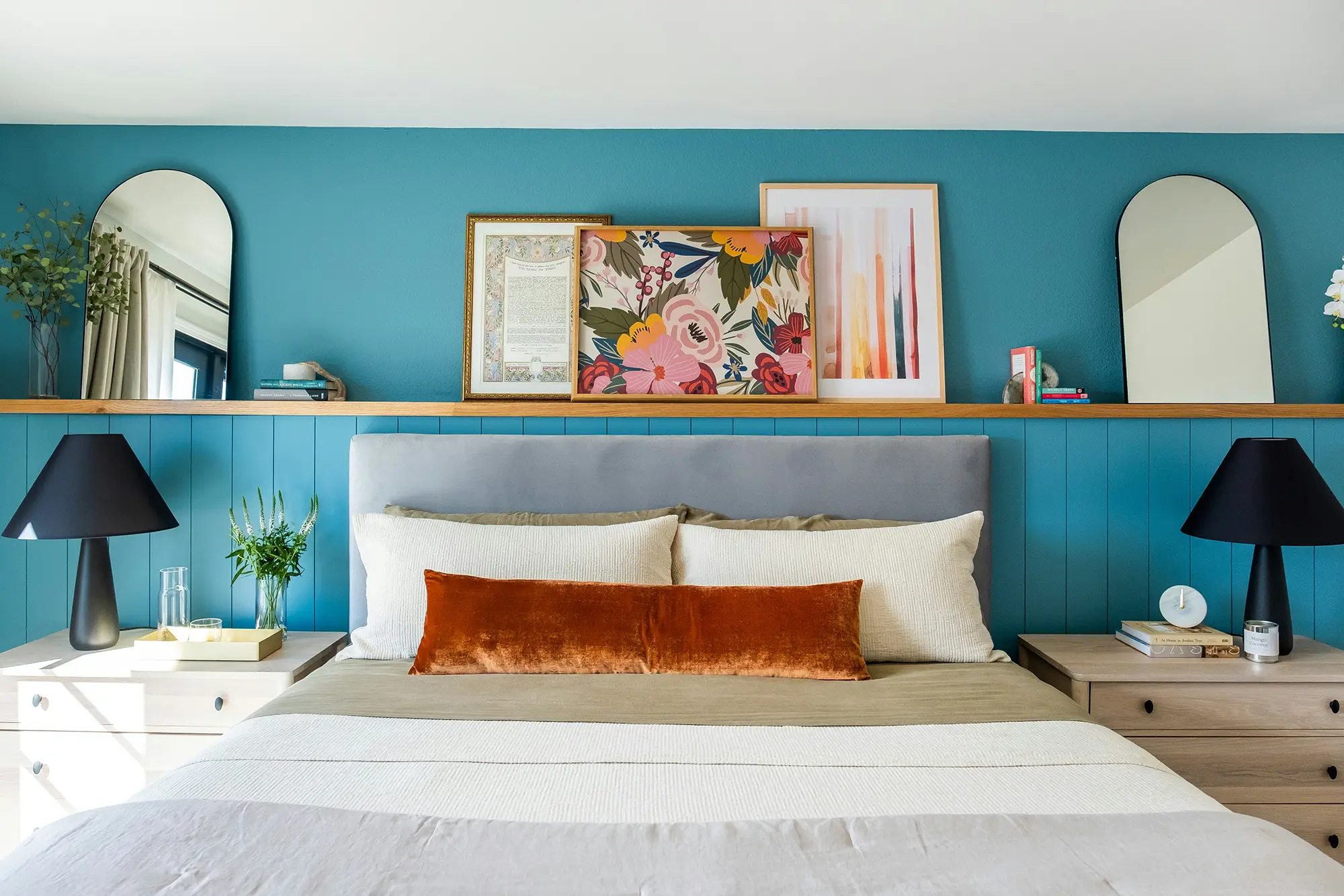 A neatly made bed with neutral bedding, orange accent pillow, nightstands with lamps, artwork and decor on a shelf against a teal paneled wall.